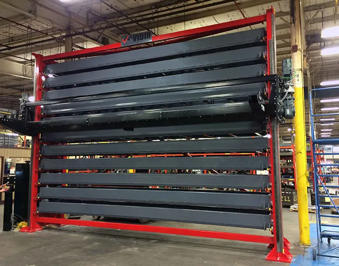A large industrial vertical lift module with multiple horizontal shelves, enclosed in a red frame, stands in a spacious warehouse. The Vidir Vertical Lift for Bar Stock Storage streamlines manufacturing efficiency, featuring dark panels surrounded by various tools and equipment.