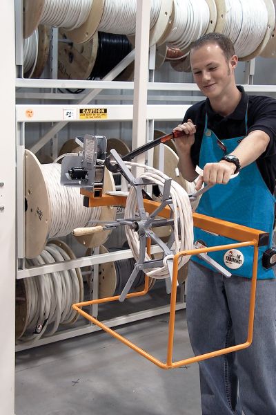A person in a blue apron is operating a manual cable coiling machine, winding white cable around it. Large spools of white cable rest on shelves in the industrial workshop, which is enhanced by prioritizing safety features to ensure a secure work environment.