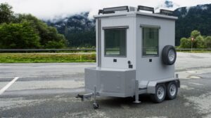 A mobile guard booth with wheels and windows stands on a paved area near a road. Its equipped with a mounted spare tire and surrounded by lush green trees and distant mountains under a cloudy sky, offering flexible security solutions in any environment.