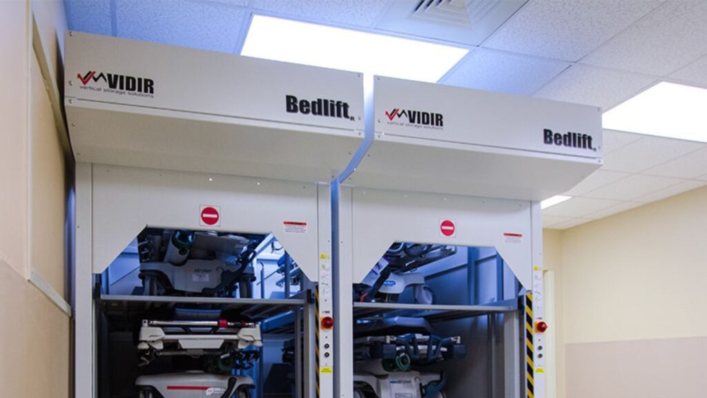 The Vidir Hospital Bedlifts, featuring customizable vertical bed storage systems, are elegantly installed in a room with a tiled ceiling and beige walls. This Bedlift efficiently stores hospital beds in multiple compartments for optimal organization.