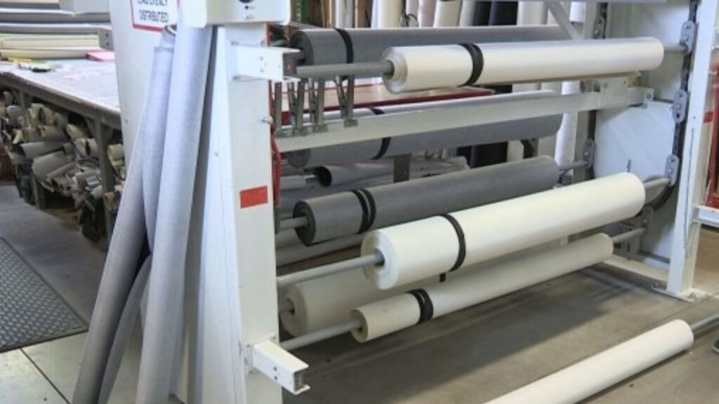 In a bustling textile workshop, rolls of fabric in shades of white and gray are neatly arranged on a metal rack. The background reveals tables and additional rolls, highlighting efficient fabric storage solutions in this busy production environment.