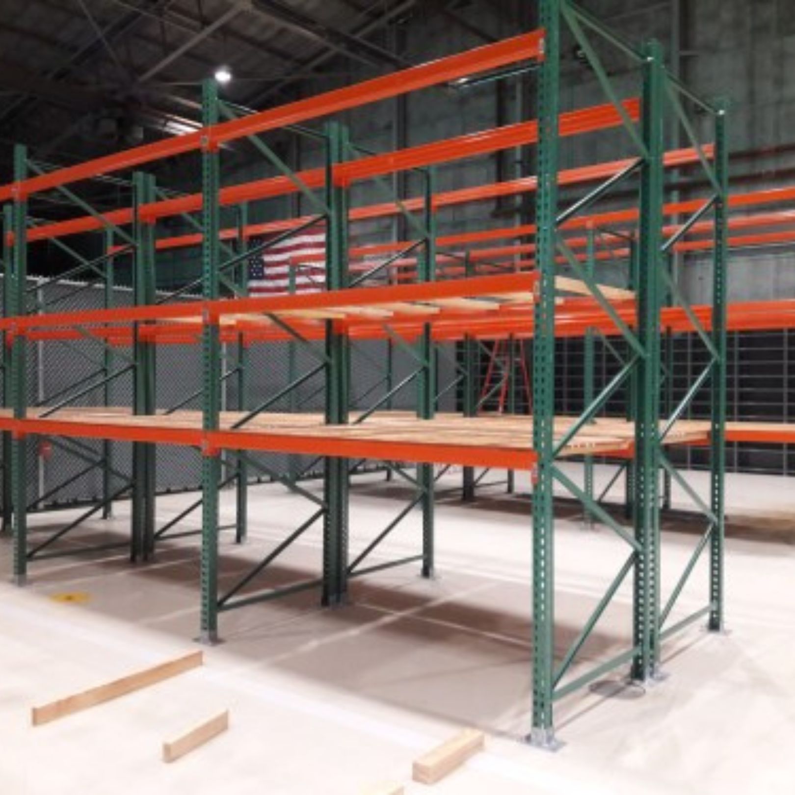 Empty warehouse shelving units with green vertical supports and orange horizontal beams, resembling oversized lockers. The concrete floor is clean, and an American flag is visible on the wall in the background.