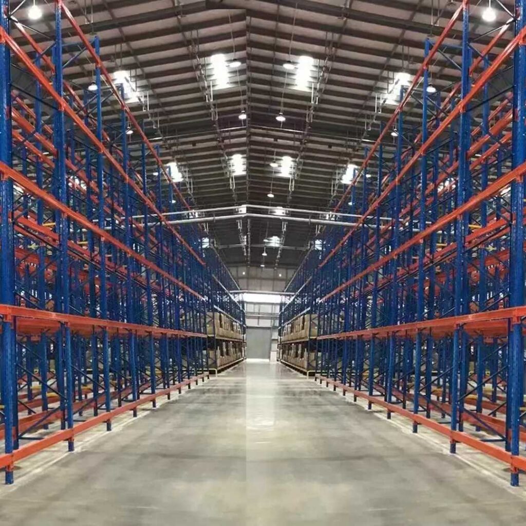 A wide-angle view of an empty warehouse showcases high, blue and orange heavy-duty racks on both sides, converging toward the center. The polished concrete floor reflects the industrial lights hanging from the ceiling above.