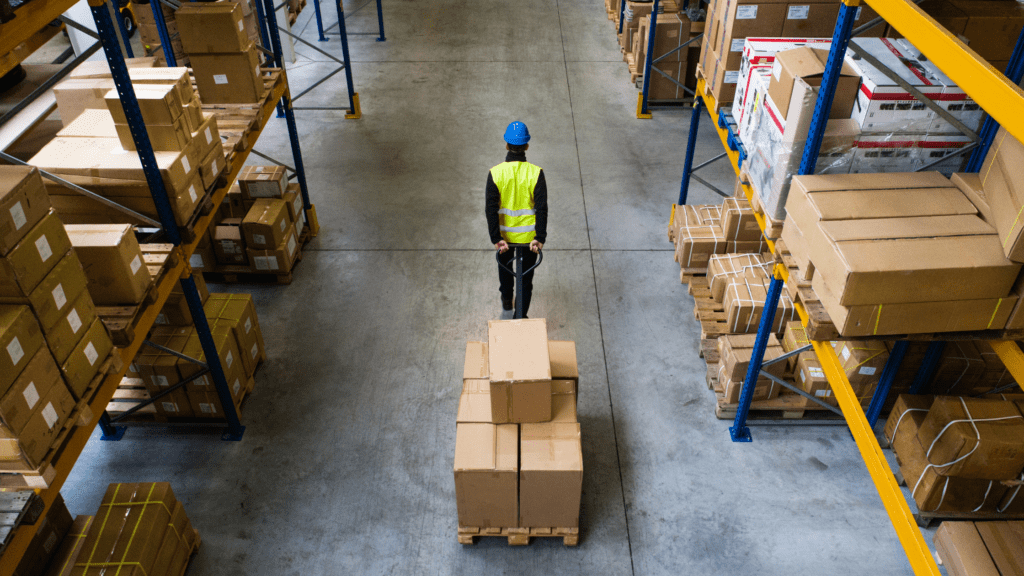 A worker in a blue helmet and yellow safety vest maneuvers a hand pallet truck loaded with boxes through an industrial warehouse, flanked by shelves showcasing smart storage ideas for moving companies. The concrete floor stretches beneath the neatly stacked cardboard boxes.