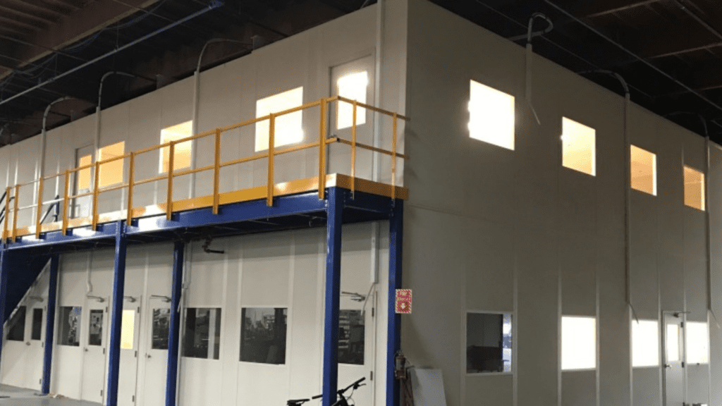 A large industrial structure with a high ceiling features an elevated platform with yellow railings, accessed by stairs. The prefabricated office building has several windows letting in light and a few doors at the lower level.