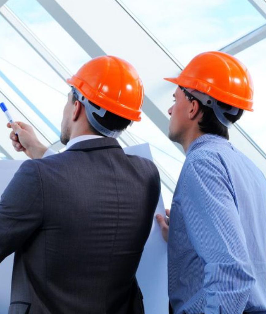 Two men in orange hard hats review building plans, with one pointing a pen. They stand by a large window, seemingly in a modern construction area, discussing Utah pallet rack regulations to ensure compliance with safety standards.