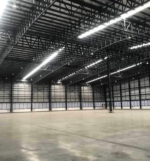 The expansive industrial warehouse features towering ceilings, metal beams, and sturdy concrete floors. Rows of bright overhead lights ensure clear visibility. Metal panel-lined walls and high windows complement its design, while adherence to OSHA warehouse guidelines ensures a safe working environment.