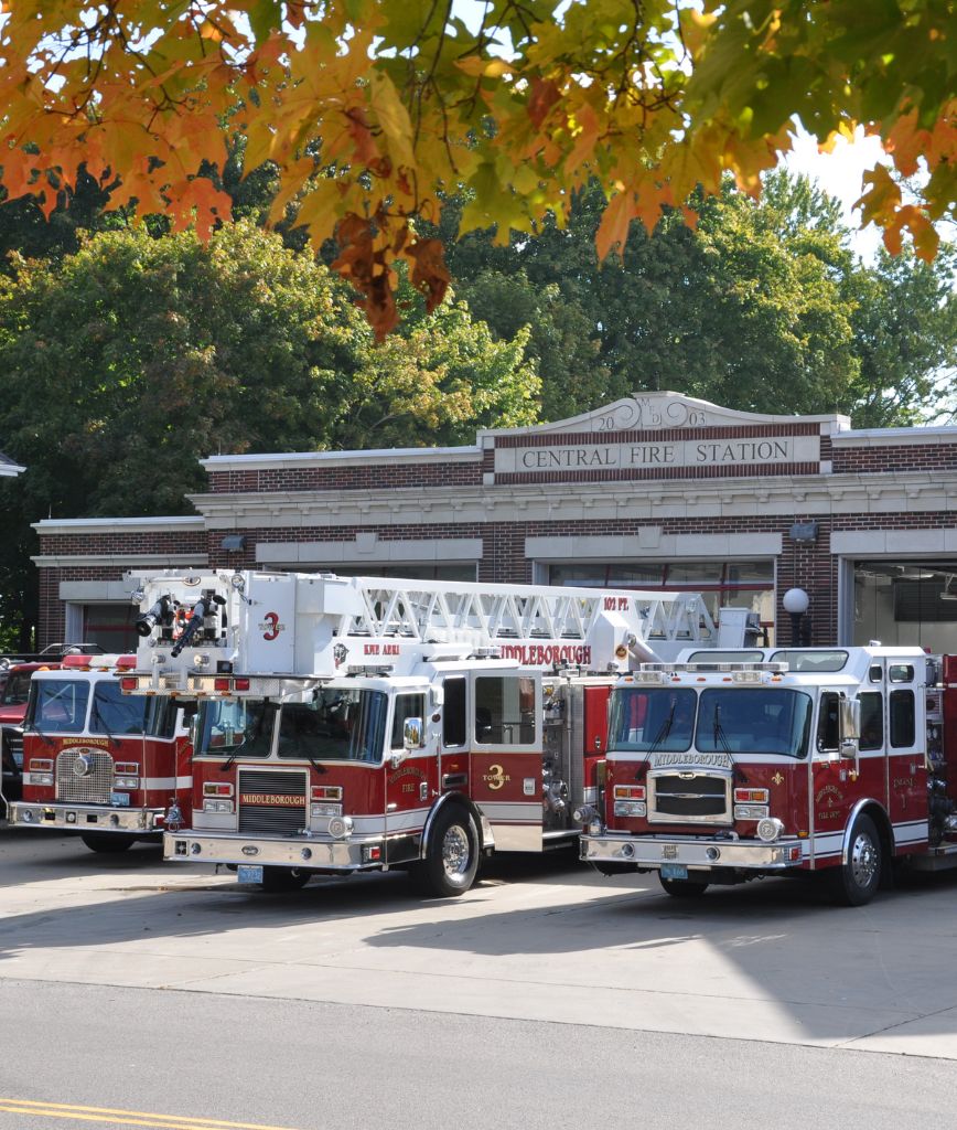 Three red fire trucks are parked outside a brick Central Fire Station, surrounded by autumn leaves and trees. In line with Utah Rack Permitting Service, ensuring prompt readiness is as crucial as meeting emergency service standards.