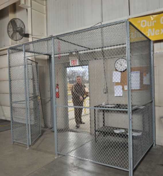A person stands at the entrance of a small room made of chain-link fencing inside a large building. The room, dedicated to warehouse safety, contains a desk with papers, a clock, and a corkboard. An exit sign and fire extinguisher are visible.