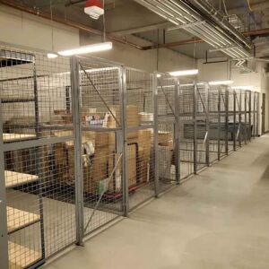 Metal storage cages containing boxes and shelves in a warehouse with concrete floors and exposed pipes on the ceiling. Fluorescent lights illuminate the area.