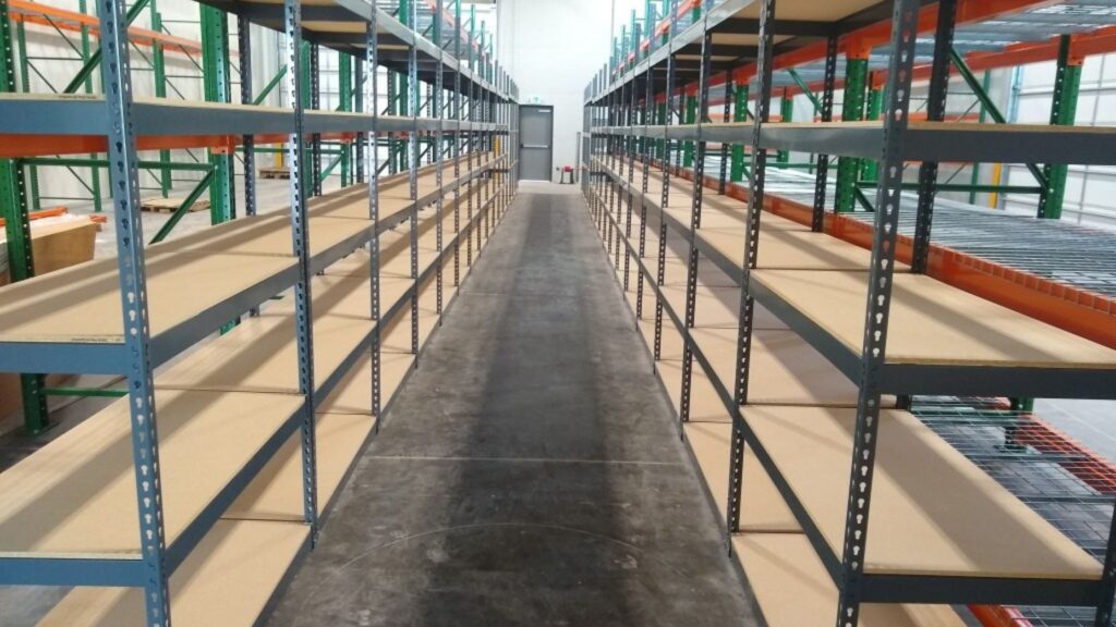 Empty industrial shelving units stand in parallel rows, offering space-efficient storage solutions in a well-lit warehouse. The metal-framed shelves with wooden panels rest on a concrete floor, while green and orange structural supports add contrast in the background.