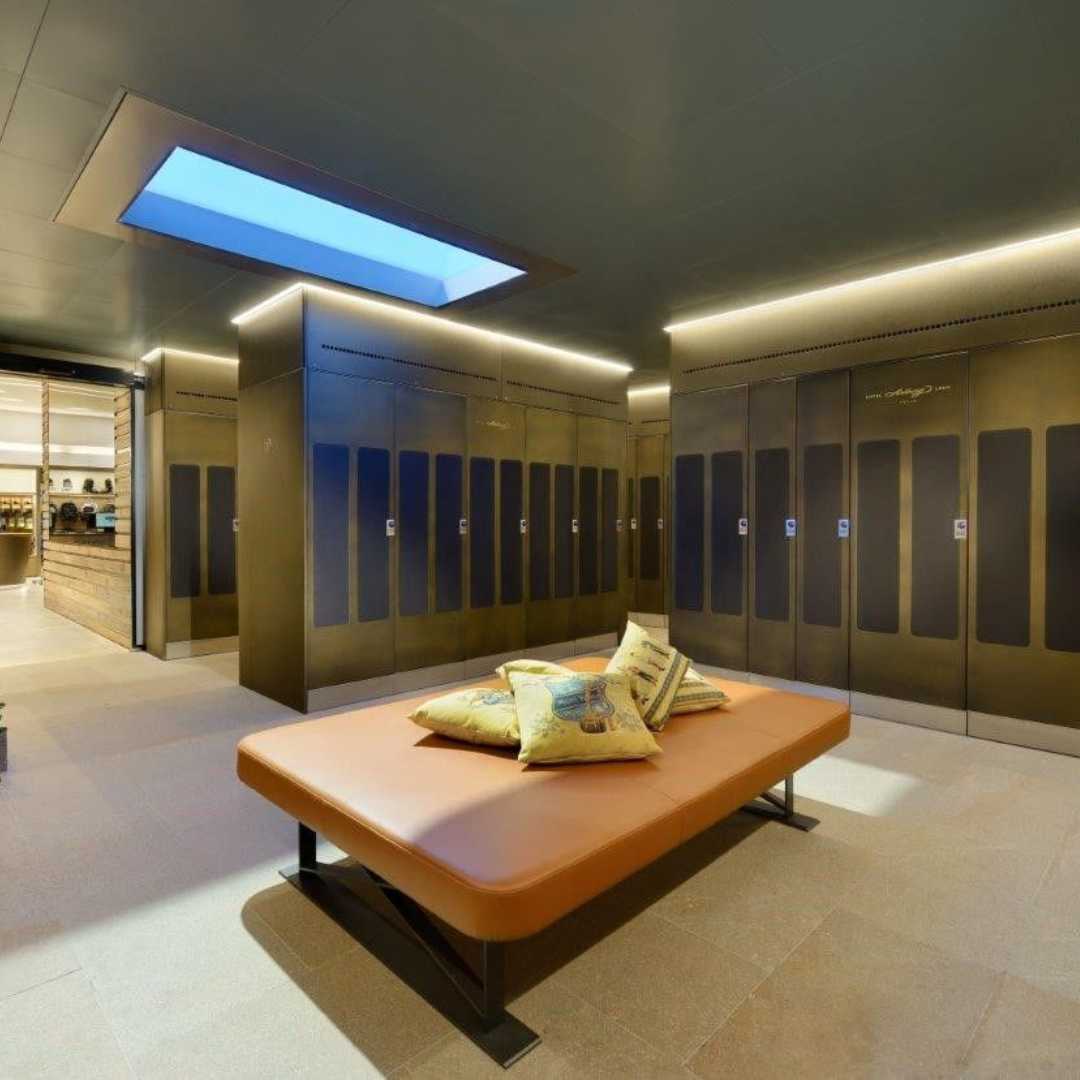 Modern locker room with sleek, dark-toned lockers, including parcel lockers, lining the walls. A central brown cushioned bench with decorative pillows resides beneath a rectangular skylight. Warm lighting illuminates the space, and a wooden counter is visible in the background.