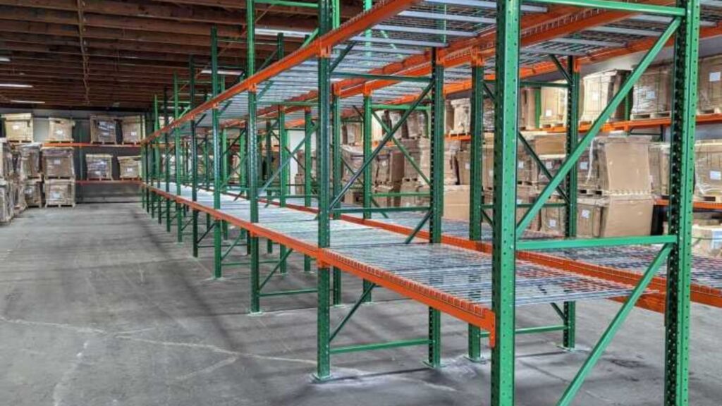 Empty green and orange industrial shelving units in a warehouse offer space-efficient storage, with stacks of boxes on pallets in the background. The concrete floor and wooden ceiling are visible, suggesting a spacious storage area.