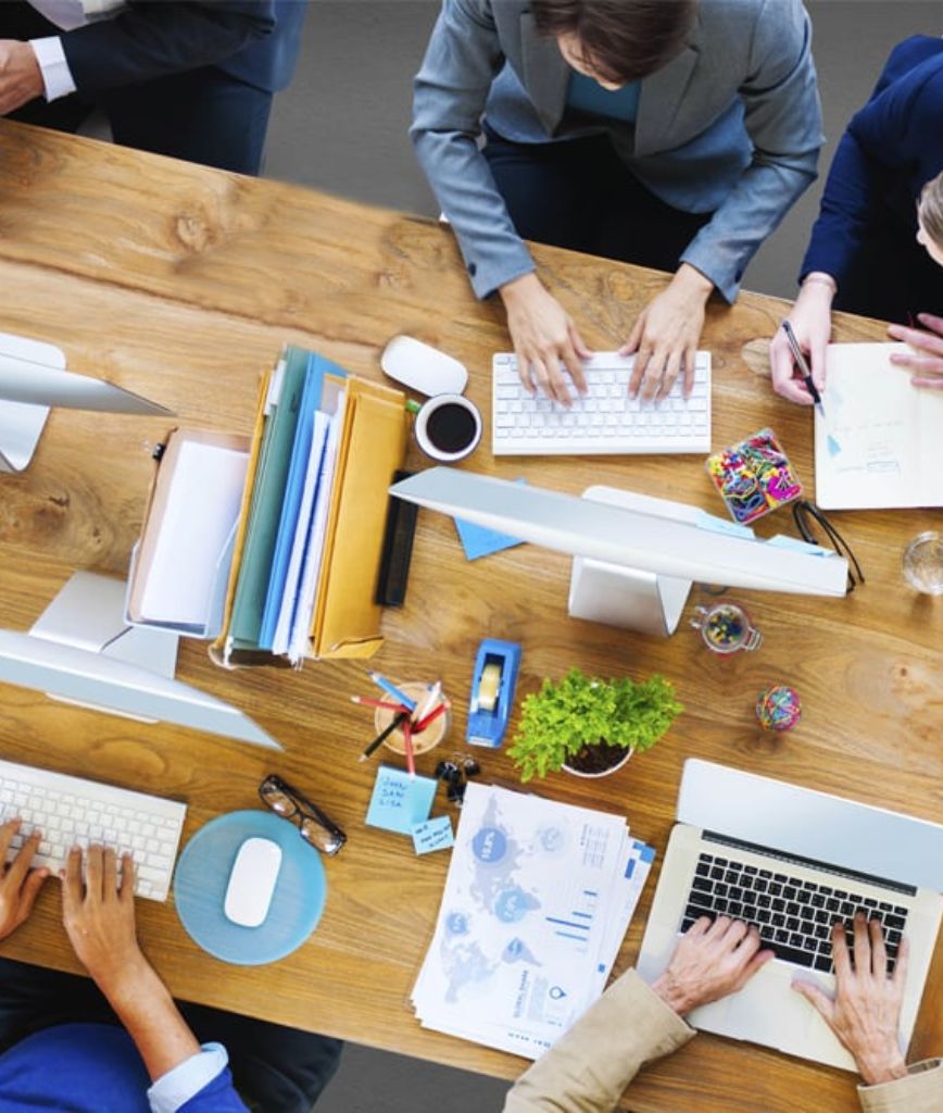 Aerial view of a collaborative workspace with four people working at a wooden table. Laptops, documents, pens, a plant, and colorful office supplies are spread across the table—like a hub for brainstorming Utah rack permitting service strategies.