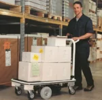 In a warehouse aisle, someone in a dark uniform smiles behind an Electro Kinetic Technologies Pony Express Motorized Platform Truck, 1500 lb. capacity, with large white boxes on its 40 L x 25 1/2 W deck. Shelves filled with more boxes highlight the trucks efficiency.
