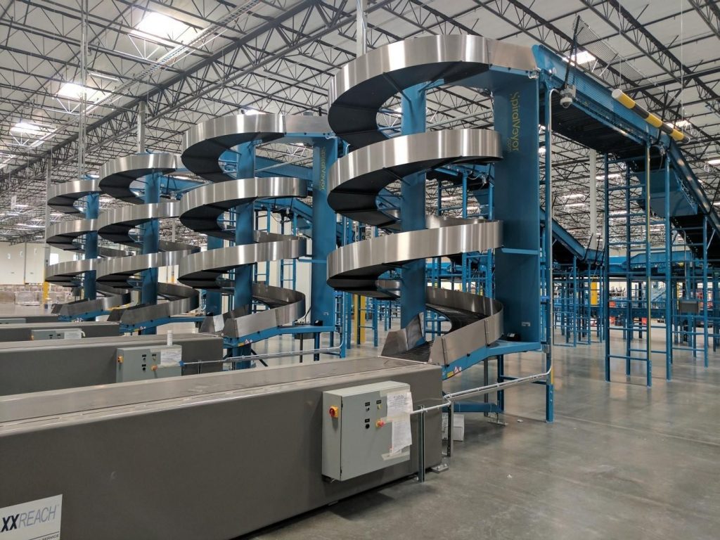 A large industrial warehouse features two curved conveyor systems. The spiral conveyors are blue and silver, extending upward. The open space around them shows high ceilings and a network of beams.