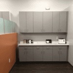 A kitchenette with gray cabinets and drawers against a light gray wall, exuding a serene healthcare-inspired ambiance. An orange accent wall is on the left, adding warmth. The countertop holds kitchen appliances and essentials, while the dark wood floor anchors the soothing space.