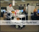 A man steers an Electro Kinetic Technologies Pony Express Motorized Mail Room Cart, able to hold up to 1500 lbs. with its baskets, through a cubicle-filled office under fluorescent lights. Two people work nearby; one at a computer and the other on the phone.