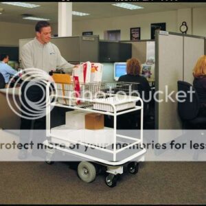A man steers an Electro Kinetic Technologies Pony Express Motorized Mail Room Cart, able to hold up to 1500 lbs. with its baskets, through a cubicle-filled office under fluorescent lights. Two people work nearby; one at a computer and the other on the phone.