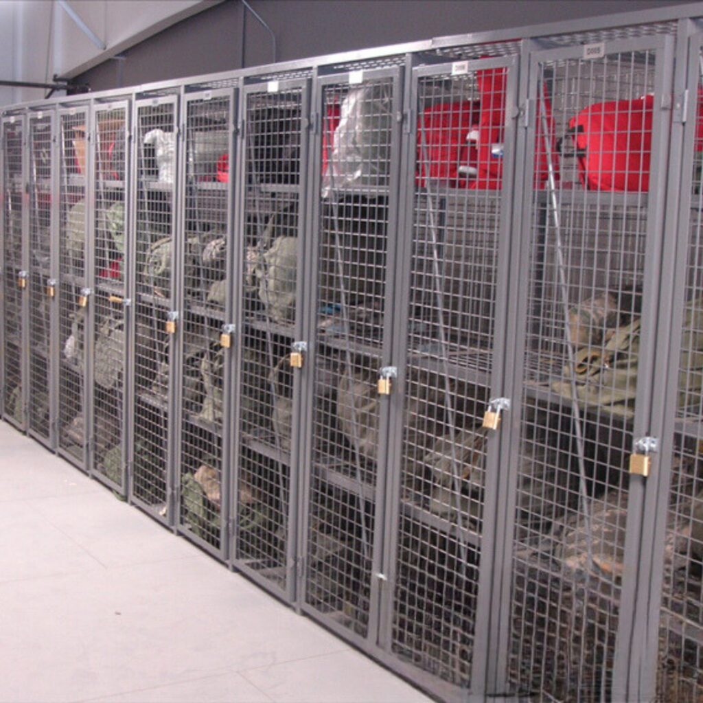 A row of metal storage lockers with mesh doors, each secured with padlocks, lines the Cogan storage area. Red bins and various items, including bags and camouflage gear, are visible inside this well-organized space.
