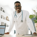 A smiling healthcare professional in a white coat and stethoscope leans forward on a desk with a laptop, papers, and a potted plant. The background features shelves adorned with personal photos and decor.