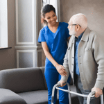 A caregiver in blue scrubs assists an elderly man using a walker, embodying true healthcare dedication. They stand in a well-lit room with a sofa and large windows, both smiling warmly and conveying genuine care and support.