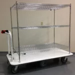 The Electro Kinetic Technologies Pony Express Motorized Stock Cart has a 1500 lb. capacity and features a 24 x 36 x 54 shelving unit with three wire shelves. It glides on wheels across the gray floor with ease, thanks to its sturdy white base and convenient handle for maneuvering.