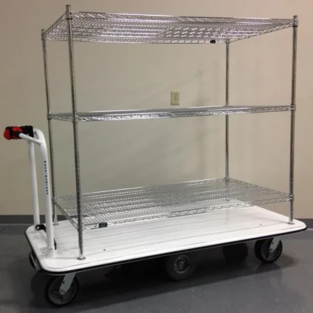 The Electro Kinetic Technologies Pony Express Motorized Stock Cart has a 1500 lb. capacity and features a 24 x 36 x 54 shelving unit with three wire shelves. It glides on wheels across the gray floor with ease, thanks to its sturdy white base and convenient handle for maneuvering.