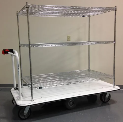 The Electro Kinetic Technologies Pony Express Motorized Stock Cart has a 1500 lb. capacity and features a 24 x 36 x 54 shelving unit with three wire shelves. It glides on wheels across the gray floor with ease, thanks to its sturdy white base and convenient handle for maneuvering.