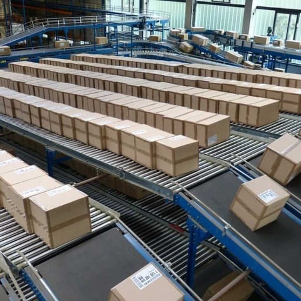 Rows of cardboard boxes travel on an AmbaFlex Conveyors system in a bustling warehouse setting, indicating a sorting or packaging process. The setup features blue supportive structures and overhead lighting, ensuring efficiency and precision.
