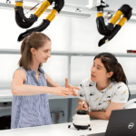 In a lab setting, embodying the spirit of healthcare research, two women engage in discussion. One, with long hair in a ponytail and a striped sleeveless shirt, gestures animatedly to the other, who listens attentively beside a microscope and laptop. Overhead hang yellow and black ventilation tubes.