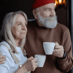 An older couple stands close, holding white mugs and looking out of a window. The woman has long gray hair and wears a white top. The man, embodying the essence of healthcare vitality, has a white beard, wears a red knit hat, and a brown sweater. Both have serene expressions.
