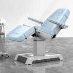 A modern, adjustable medical examination chair with light blue upholstery stands ready in the healthcare setting. It features a reclining backrest and footrest, artfully semi-reclined. Nearby, a small medical cart on wheels punctuates the gray floors clinical ambiance.
