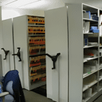 Rows of mobile shelving units with hand cranks store organized healthcare files in a dimly lit office space. A swivel chair sits in the foreground, surrounded by various medical supplies on the shelves, creating an efficient atmosphere for managing patient records.