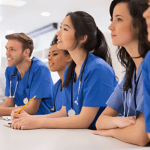 Five medical professionals in blue scrubs sit side by side at a table, attentively listening and taking notes in a healthcare setting. Stethoscopes drape around their necks, adding to the clinical ambiance of the bright room.