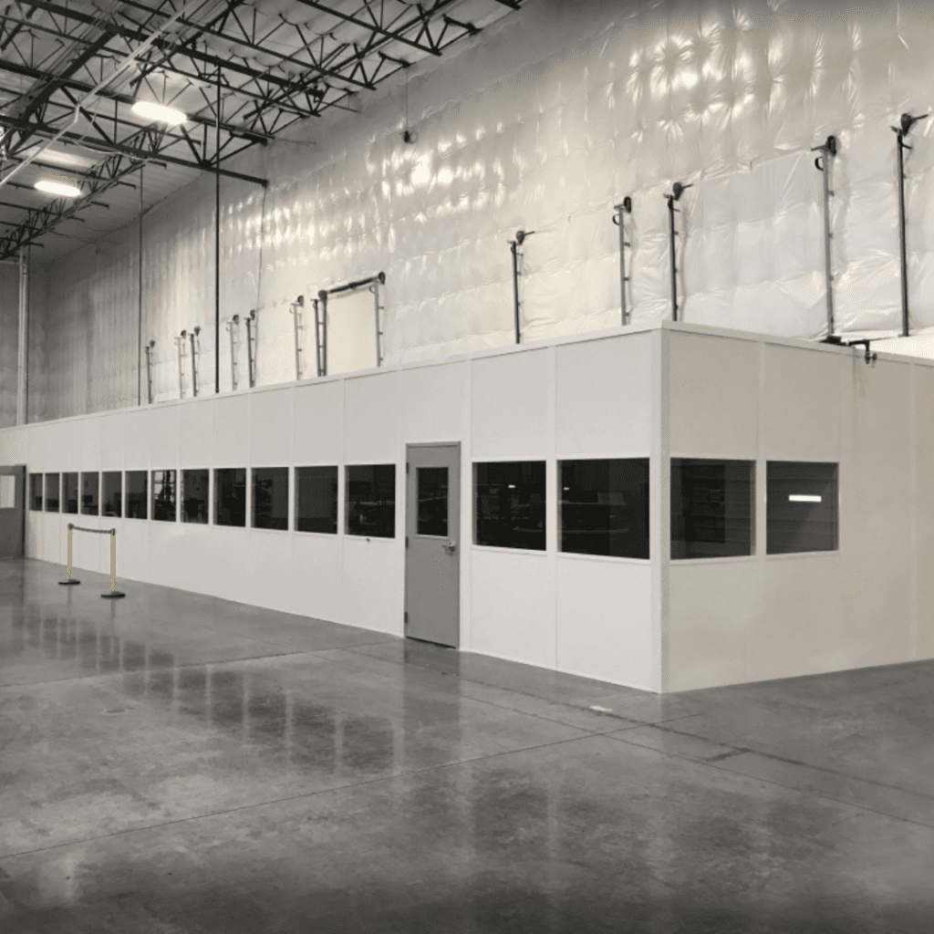 Industrial warehouse interior with a large, white, enclosed office space featuring multiple dark-tinted windows and a metallic finish. Ideal for healthcare logistics, the space has visible ceiling tiles and lighting fixtures, plus a yellow safety barrier for added protection.