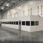 Industrial warehouse interior with a large, white, enclosed office space featuring multiple dark-tinted windows and a metallic finish. Ideal for healthcare logistics, the space has visible ceiling tiles and lighting fixtures, plus a yellow safety barrier for added protection.