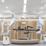 A modern healthcare operating room features a surgical table at its center, surrounded by overhead lights, medical monitors, and equipment on mounted arms. The room maintains a clean, sterile appearance with beige curtains in the background.