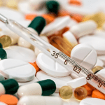 A close-up of various pills, including white tablets and colorful capsules, scattered around a glass thermometer. This healthcare scene features a thermometer displaying a temperature scale.