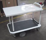 A white Electro Kinetic Technologies Pony Express Motorized Platform Truck, boasting a 1500 lb. capacity and (2) shelving units of 26 wide x 40 long, is parked in an industrial setting with marked yellow guidelines on the concrete floor. It features a handlebar and four wheels.