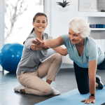 An older woman in a light blue shirt balances on her hands and knee, extending one arm and leg—a testament to her strength. A younger woman in a gray shirt kneels beside her, offering healthcare support. In the background, a blue exercise ball adds to the fitness scene.