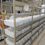Metal shelves in the warehouse department hold rows of white cardboard boxes. A clear plastic bag labeled Document Enclosed hangs on the top shelf, while more boxes and shelves stretch into the spacious storage area.