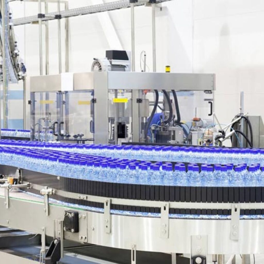 Bottling plant conveyor line with clear plastic water bottles and blue caps. The bottles glide smoothly along the AmbaFlex Conveyors metal track, passing through machinery in a well-lit industrial setting.
