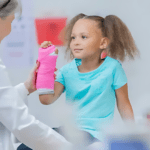 A young girl in a turquoise shirt sits and smiles, her arm in a pink cast. In the supportive environment of a healthcare office, a person in a white coat gently holds her hand, providing reassurance and care.