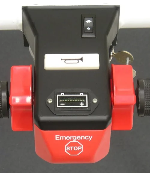 Close-up of a red and black emergency stop device from the Electro Kinetic Technologies Pony Express Motorized Platform Truck, 1500 lb. capacity (40 L x 25.5 W deck), featuring a red Emergency STOP button, keyhole, bell icon switch, and connector port for enhanced safety on any platform. Background is gray.