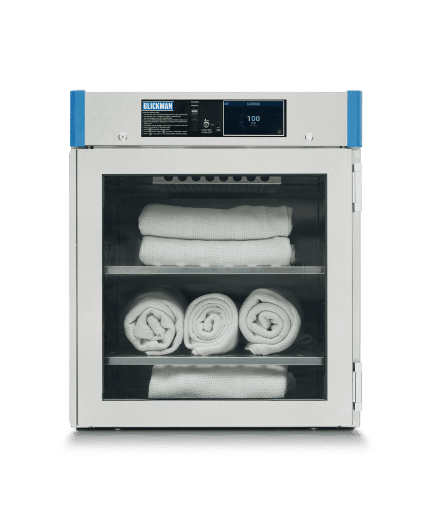 Front view of a Blickman white and blue sterilizer cabinet with a glass door. Inside, neatly stacked, rolled white towels rest on two shelves. The digital display shows a temperature reading, and labels are visible on the top front.