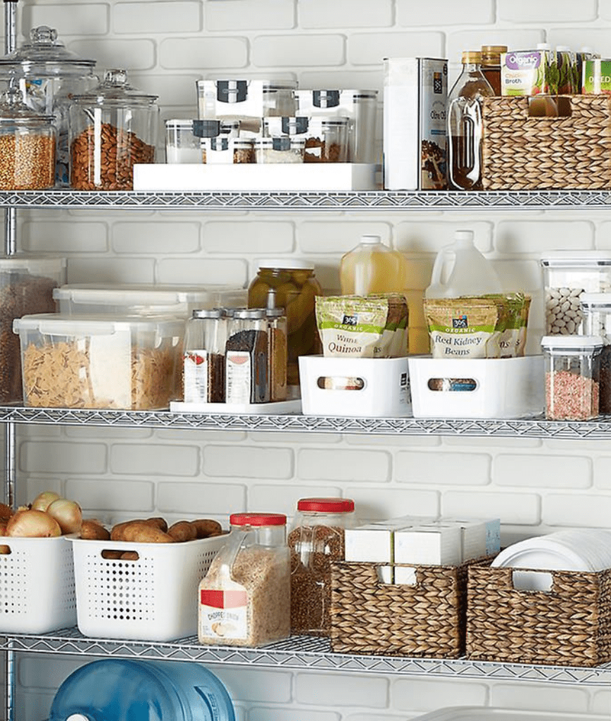 A well-organized pantry with wire shelves holding various items. Glass jars and baskets contain grains and pasta. Plastic bins hold food items like potatoes and onions. Bottles and boxes are neatly arranged against a white brick wall.
