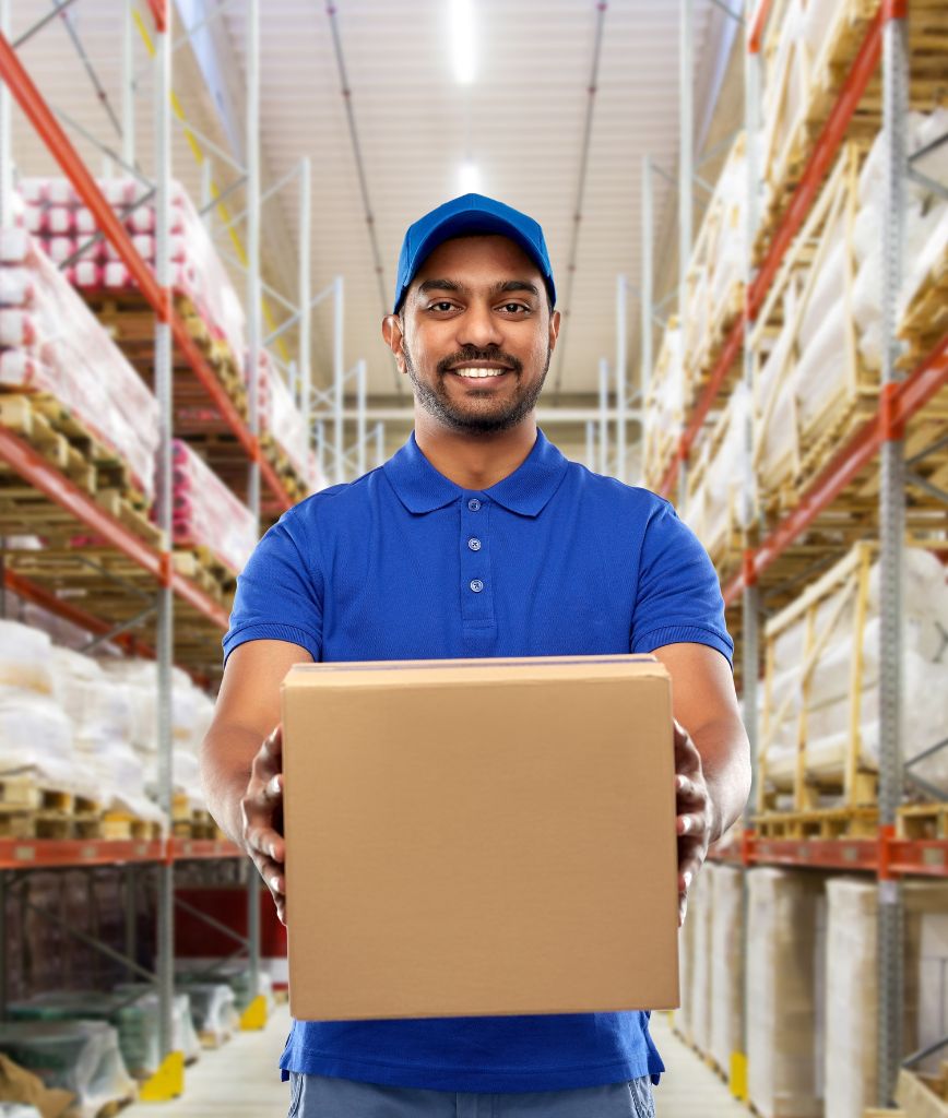 A smiling man in a blue uniform and cap holds a cardboard box in a warehouse aisle, showcasing seamless material handling solutions. The aisle is lined with shelves brimming with packages, reflecting the efficiency of warehouse automation for next-day delivery under bright lights and high ceilings.