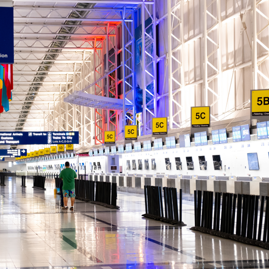 An empty airport check-in area with rows of unoccupied counters and a swab booth for virus testing stands ready. The ceiling and walls are illuminated in red, blue, and white lights. A couple of travelers linger to the left, while floor signs display gate numbers.