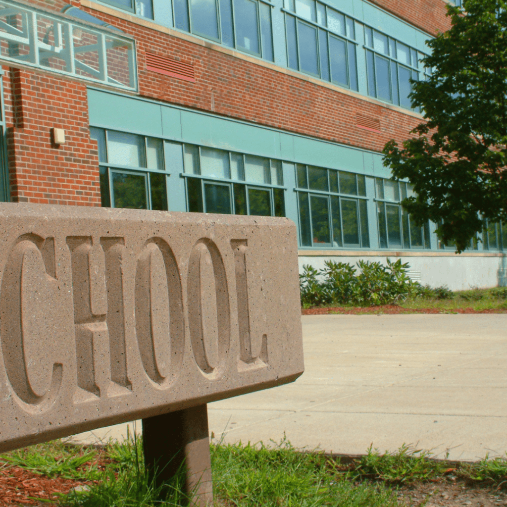 A stone sign reading SCHOOL stands in front of a red brick building with large windows. Sparse greenery surrounds the area, which now features a swab booth for virus testing under the clear sky, ensuring health and safety for all.